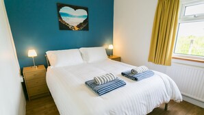 Un lit bien fait, avec du linge de lit blanc et des serviettes à rayures, un mur bleu orné d’une œuvre d’art en forme de cœur, et une fenêtre avec des rideaux jaunes.