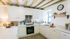 A modern kitchen with white cabinets, a built-in oven, a Bosch dishwasher, wooden flooring, and a clock on the wall. - Ysgubor Wennol (Swallow Barn) -  a cosy fire that sleeps 4 guests  in 2 bedrooms (Llanfairpwll)