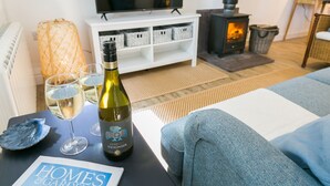 A cozy living room with a sofa, a table with a bottle of wine and glasses, a TV, a fireplace, and storage baskets. - Ysgubor Wennol (Swallow Barn) -  a cosy fire that sleeps 4 guests  in 2 bedrooms (Llanfairpwll)