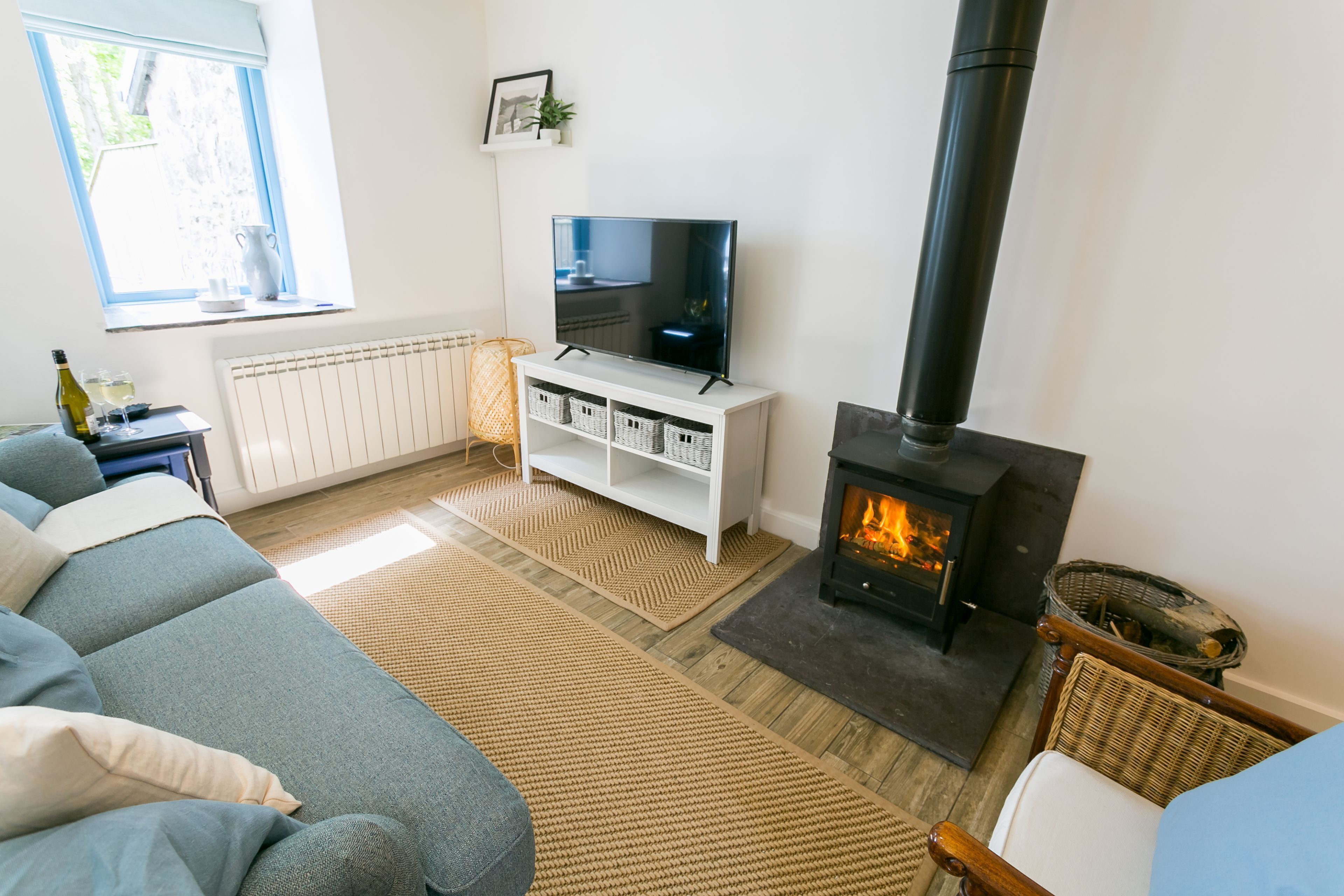Een woonkamer met een bank, een televisie op een witte dressoir, een open haard en een raam met uitzicht op bomen.