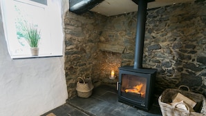 A stone-walled room with a wood-burning stove, a window with a plant, and a woven basket on the floor. - Bryn Eithin -  a remote that sleeps 2 guests  in 1 bedroom (Bodorgan)