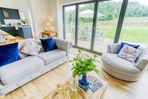 A modern living room with a grey sofa, blue cushions, a coffee table with a plant, and a view of a garden through large sliding doors.
