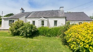 Une maison blanche à deux étages, surmontée d’un toit gris, entourée d’une végétation luxuriante et d’un arbuste jaune en fleurs.