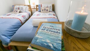 Une chambre à coucher avec deux lits, une bougie allumée et un livre intitulé « L’invention des nuages » de Richard Hamblyn.