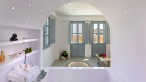 A bathroom with a bathtub, shelves with toiletries, a towel rack, and a view of the outside. - BlackStone Luxury Suites (Santorini)