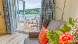 Ein Zimmer mit Blick auf einen Fluss und Grünflächen, eine Holzterrasse mit Tisch und Stühlen sowie eine Couch mit Kissen.