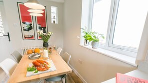 A dining area with a wooden table, chairs, a vase with flowers, a plate of food, and a window with plants.