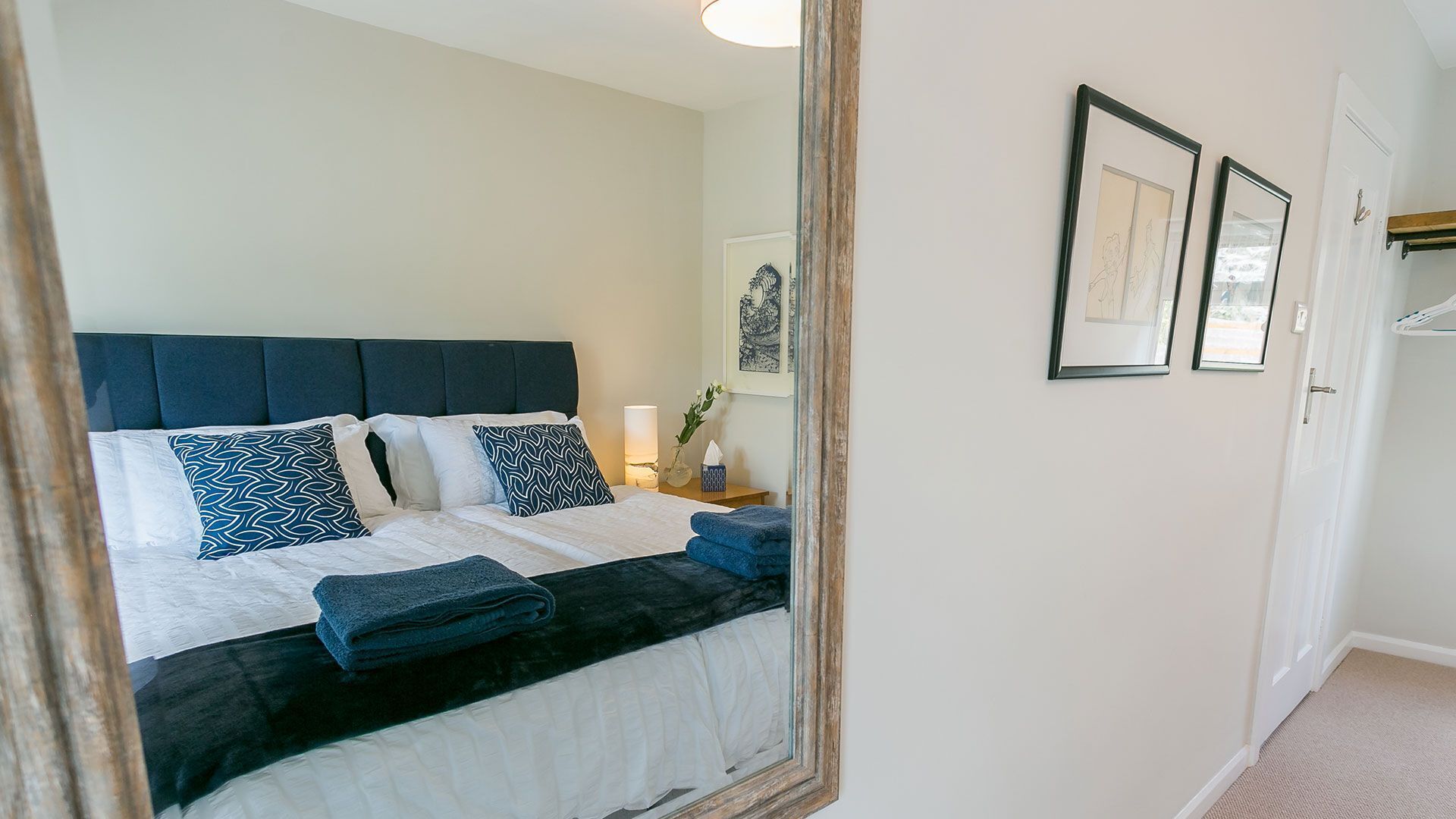 Cama bem arrumada com cabeceira escura, roupa de cama branca e almofadas estampadas. Uma mesa de cabeceira com luminária e um vaso de flores é visível. O quarto tem um design minimalista com um espelho grande refletindo a cama e uma porta com porta-toalhas.
