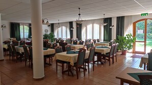 A spacious dining area with multiple tables and chairs, large windows with curtains, and potted plants. - Ágnes Hotel (Hévíz)