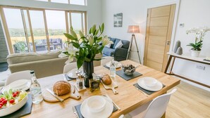 A dining area with a wooden table set for two, a vase of white flowers, and a view of the outdoors through large windows. - Bwncath & Hebog -  a coastal that sleeps 8 guests  in 4 bedrooms (Llanfairynghornwy)