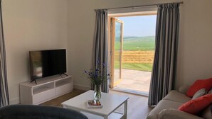 A living room with a flat-screen TV on a white stand, a coffee table with a vase of flowers, a sofa with red cushions, and a sliding glass door leading to a patio with a view of green fields. - Bwncath & Hebog -  a coastal that sleeps 8 guests  in 4 bedrooms (Llanfairynghornwy)