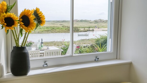 A window with a view of a lake, a boat, and a vase with sunflowers on a windowsill. - Min y Don -  an on the beach that sleeps 4 guests  in 2 bedrooms (Aberffraw)