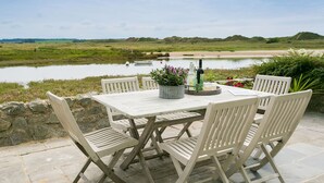 An outdoor table and chairs set on a stone patio with a view of a lake and distant hills. - Min y Don -  an on the beach that sleeps 4 guests  in 2 bedrooms (Aberffraw)