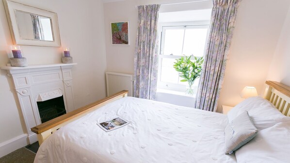 A bedroom with a white bed, a fireplace, a window with curtains, and a painting on the wall.