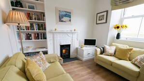 A cozy living room with a fireplace, a bookshelf, a TV, and a window with sunflowers. - Min y Don -  an on the beach that sleeps 4 guests  in 2 bedrooms (Aberffraw)
