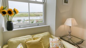 A room with a window overlooking a landscape, a sofa with cushions, and a table with a lamp. - Min y Don -  an on the beach that sleeps 4 guests  in 2 bedrooms (Aberffraw)