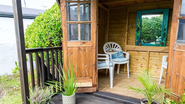 A wooden cabin with a porch, two white plastic chairs, and a small table with a cushion.