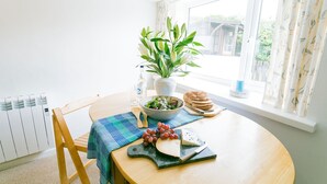 A dining table with a salad bowl, grapes, cheese, and bread, a potted plant, and a window with curtains. - Borth Bach -  an on the beach that sleeps 4 guests  in 2 bedrooms (Rhosneigr)