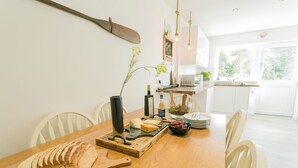 Un coin repas avec une table en bois, des chaises blanches, un vase noir contenant une plante, une planche à fromage et une bouteille de vin.