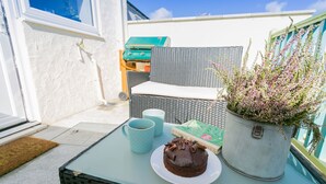 Un petit espace extérieur aménagé avec une chaise en osier, une table sur laquelle se trouvent un gâteau et deux tasses, ainsi qu’une plante en pot.