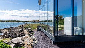 Un bâtiment moderne en verre offrant une vue sur un paysage côtier, comprenant une côte rocheuse et une étendue d’eau calme.