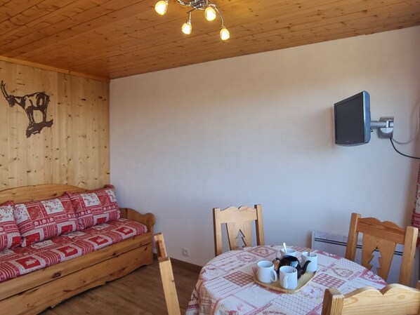 Une pièce chaleureuse avec un plafond en bois, un canapé agrémenté de coussins à motifs, une table à manger sur laquelle se trouve un plateau rempli de tasses, et un téléviseur fixé au mur.