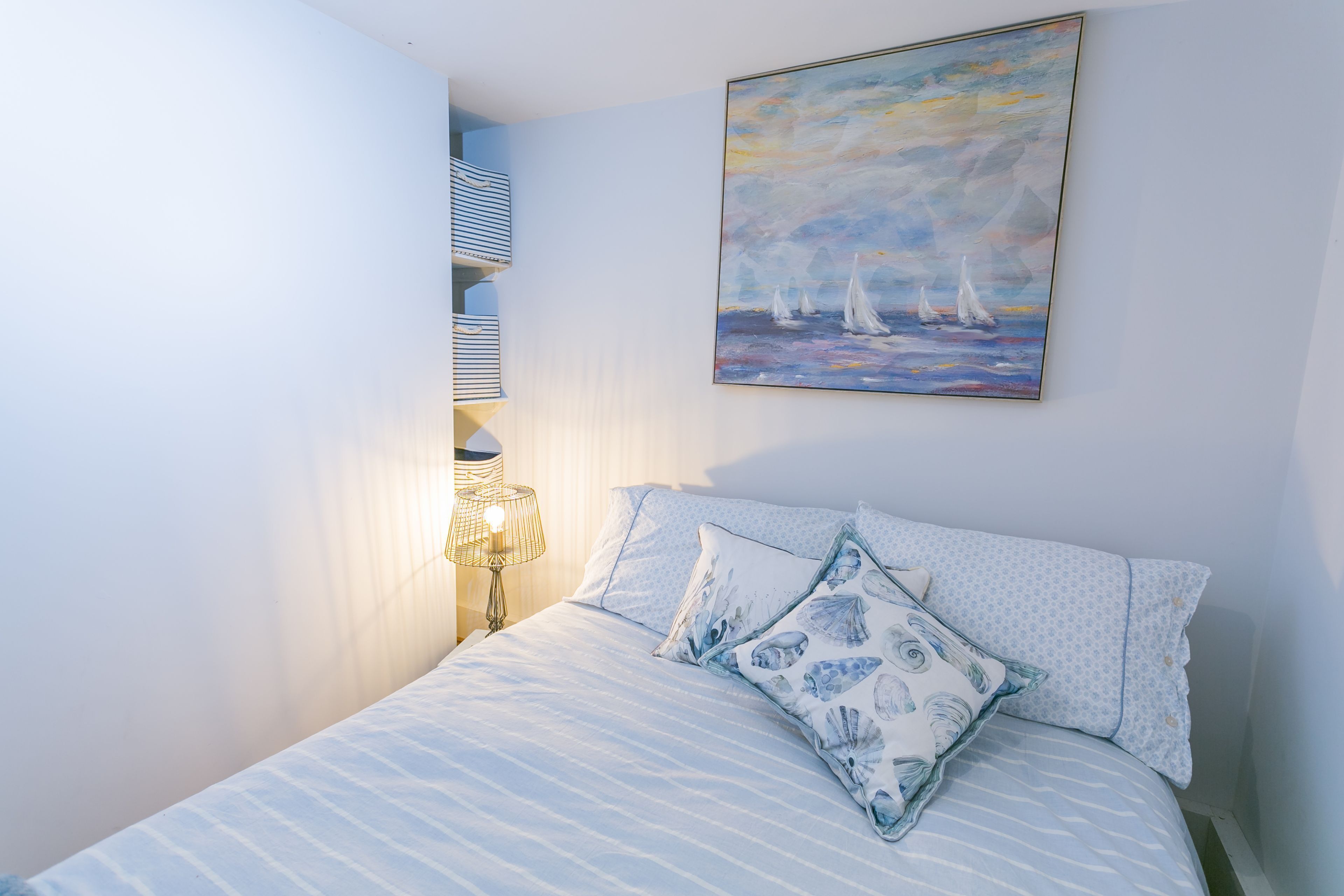 A bedroom with a bed, pillows, a nightstand, a lamp, and a painting of sailboats on the wall.