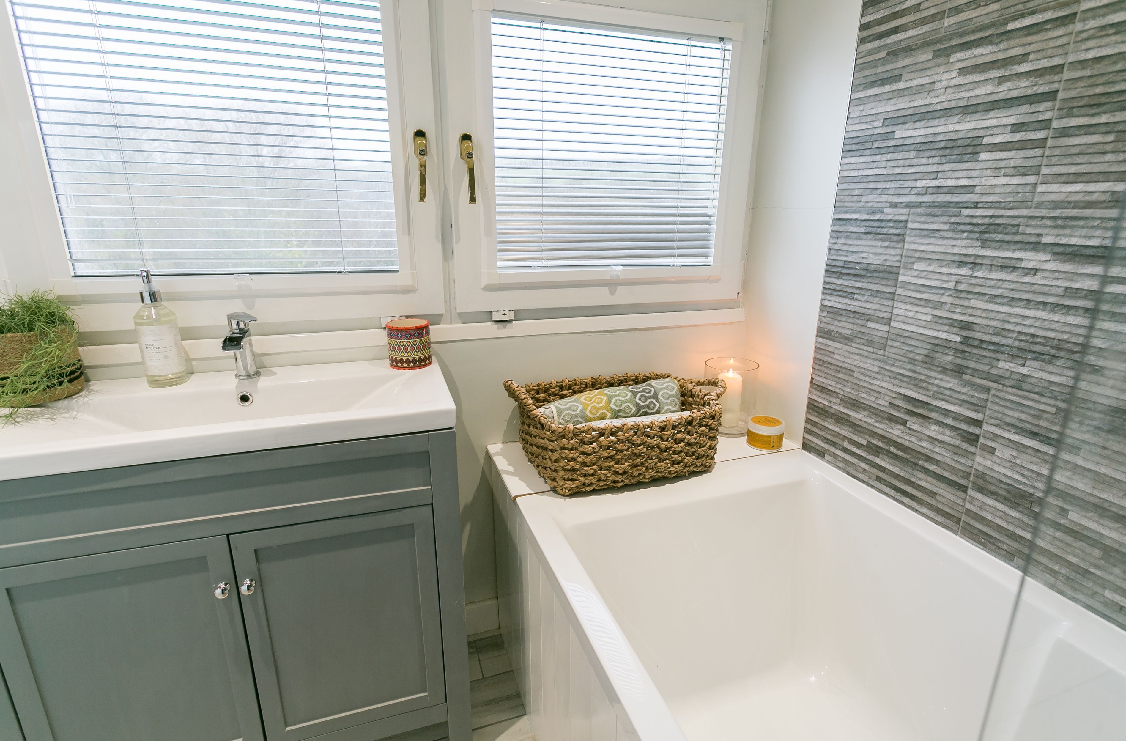 A bathroom with a white bathtub, a sink, and a window with blinds. There is a wicker basket with a cushion on the edge of the bathtub.