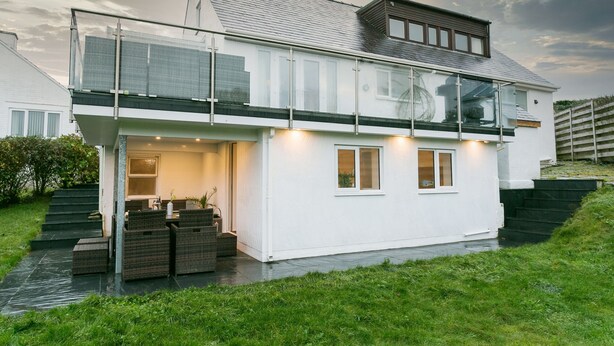 A modern two-story house with a glass balcony, a covered outdoor seating area, and a well-maintained lawn.