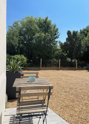 Eine Terrasse mit Holztisch und Bank, umgeben von Kies und Bäumen.
