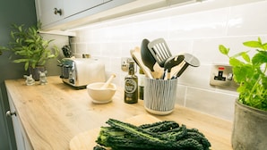 Een keuken met een houten aanrecht, een snijplank met groene bladgroenten, een broodrooster, een kom, een plant en keukengerei.