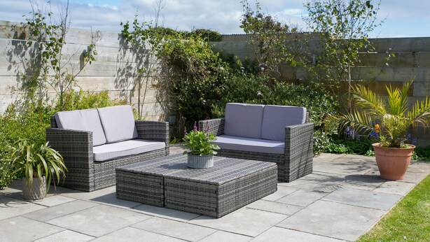 Een terras buiten met rieten meubilair, waaronder twee banken en een bijzettafeltje, omgeven door potplanten en een betonnen muur.