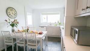 Een moderne keuken met witte kasten, een eettafel met stoelen, een klok aan de muur en een raam dat natuurlijk licht binnenlaat.