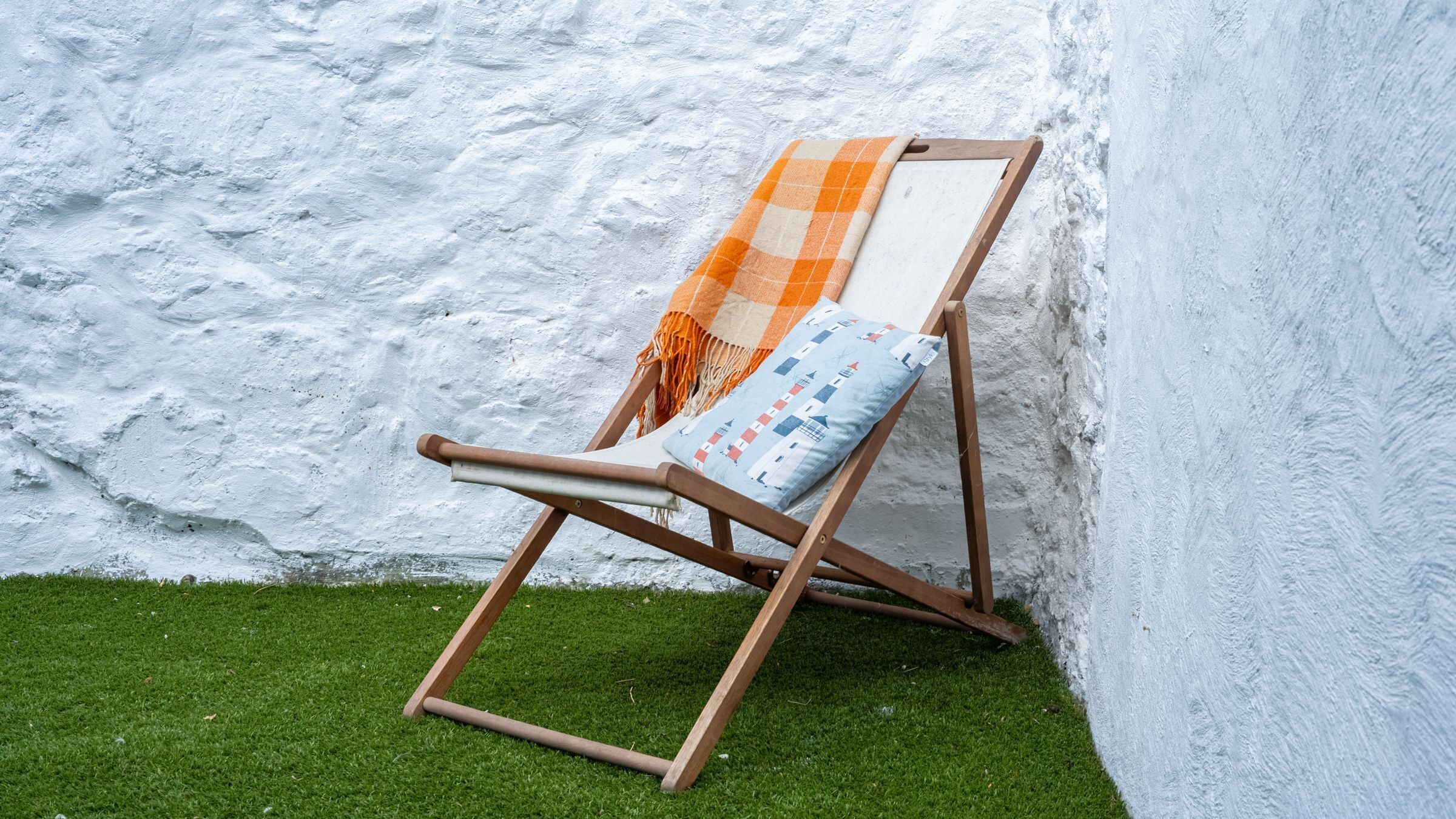 Een houten lounge stoel met een geruit kleed en een kussen, geplaatst tegen een witte muur op kunstgras.