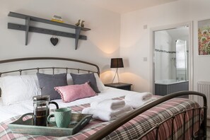Una cama bien hecha con almohadas, una mesita de noche con una lámpara, una bandeja con una taza, y un estante en la pared con libros y objetos decorativos.
