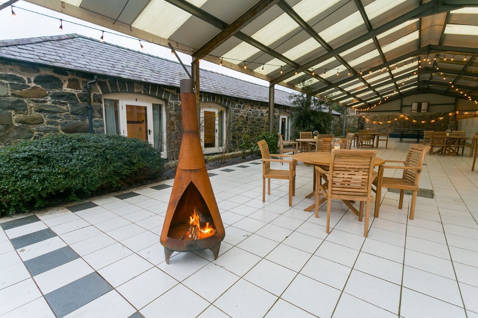 An outdoor patio with a stone building in the background, a fire pit, and wooden tables and chairs.