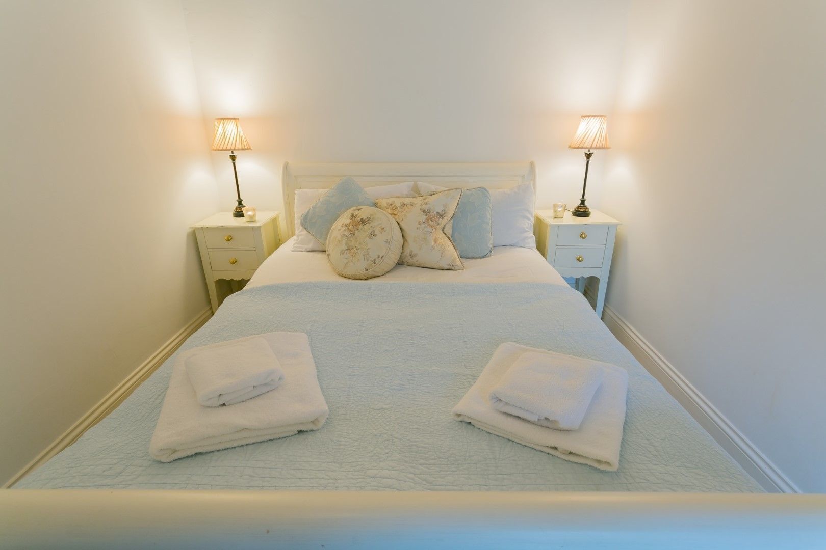 A neatly made bed with two pillows, two bedside tables with lamps, and folded towels on the bed.