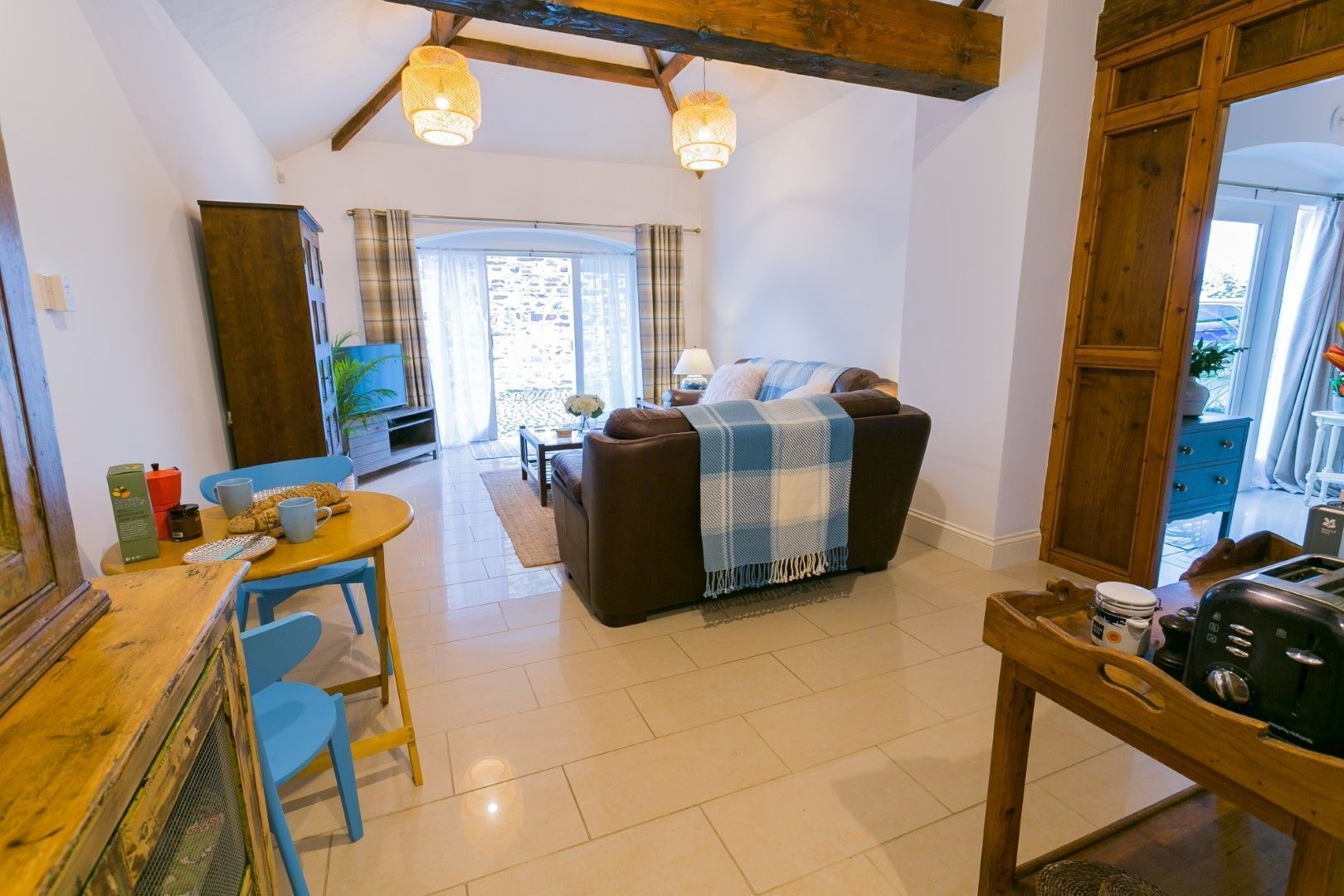 A cozy living area with a sofa, a coffee table, and a dining table with chairs. There is a wooden ceiling with exposed beams and a window with curtains.