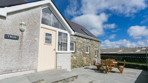A stone building with a beige door and windows, a sign reading "Y Beudy", and a patio with wooden furniture.