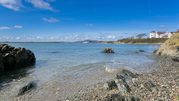 A coastal scene with clear water, pebbly beach, and a distant building.