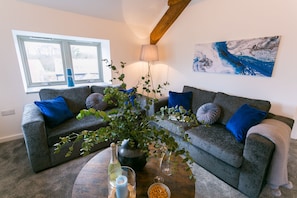 A living room with two grey sofas, blue cushions, a large plant, a wine bottle, and a painting on the wall.