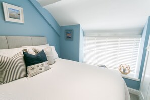 Relax in this inviting bedroom with a large bed, striped bedding, and multiple pillows, featuring a sloped ceiling and a window with white blinds.