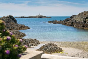 Un paisaje costero rocoso con aguas azules cristalinas, una pequeña isla con un faro, y un primer plano de algas y rocas.