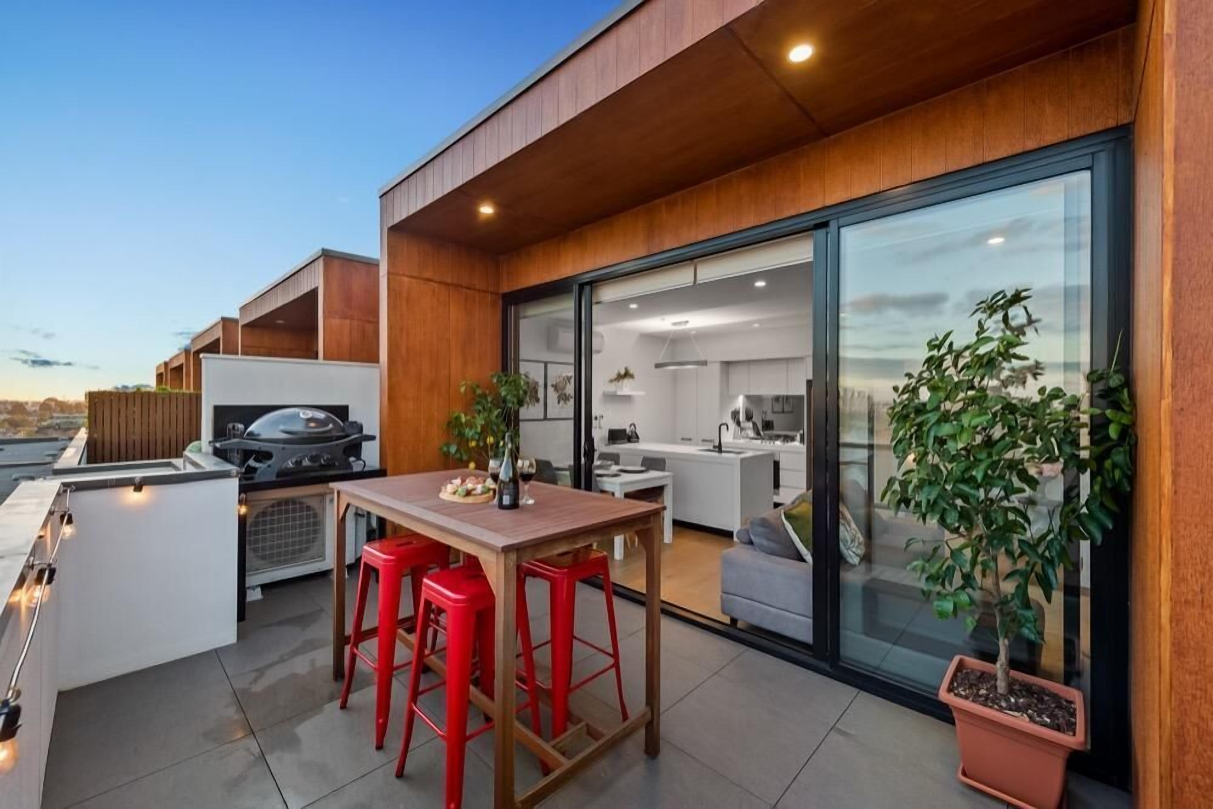 Un espace cuisine extérieur moderne, doté d’un barbecue, d’une table à manger avec des tabourets rouges et de plantes en pot.