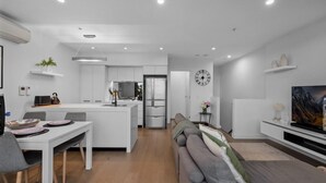 Ein modernes Wohnzimmer mit grauem Sofa, einem Esstisch mit Stühlen, einer Küchenzeile mit weißen Schr änken und einem Flachbildfernseher auf einem weißen Lowboard.