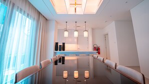 Une chambre d’hôtel moderne avec une longue table à manger, des chaises rembourrées, des luminaires suspendus et une vue sur l’extérieur à travers des rideaux transparents.