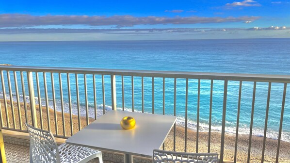 Een balkon met een tafel en stoelen met uitzicht op een strand en helderblauw water, met land in de verte.