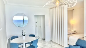 Een moderne slaapkamer met uitzicht op zee, een ronde spiegel, een rond tafeltje met stoelen, en een bed met een wit hoofdeinde.