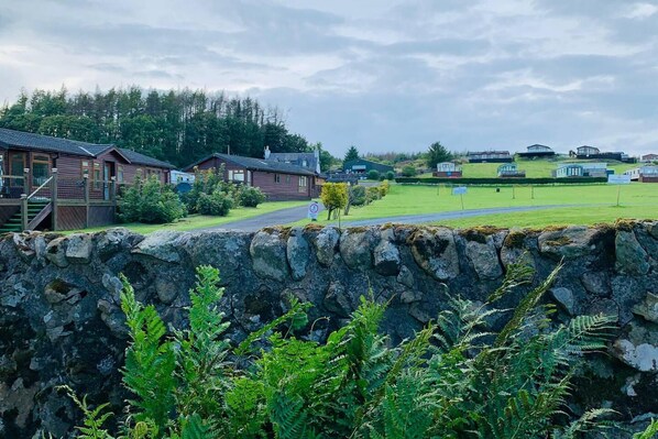 A stone wall with ferns in the foreground, a wooden building with a porch and steps, and a grassy field with several other buildings in the background. - Stunning 2-bed Apartment in Newton Stewart (Newton Stewart)