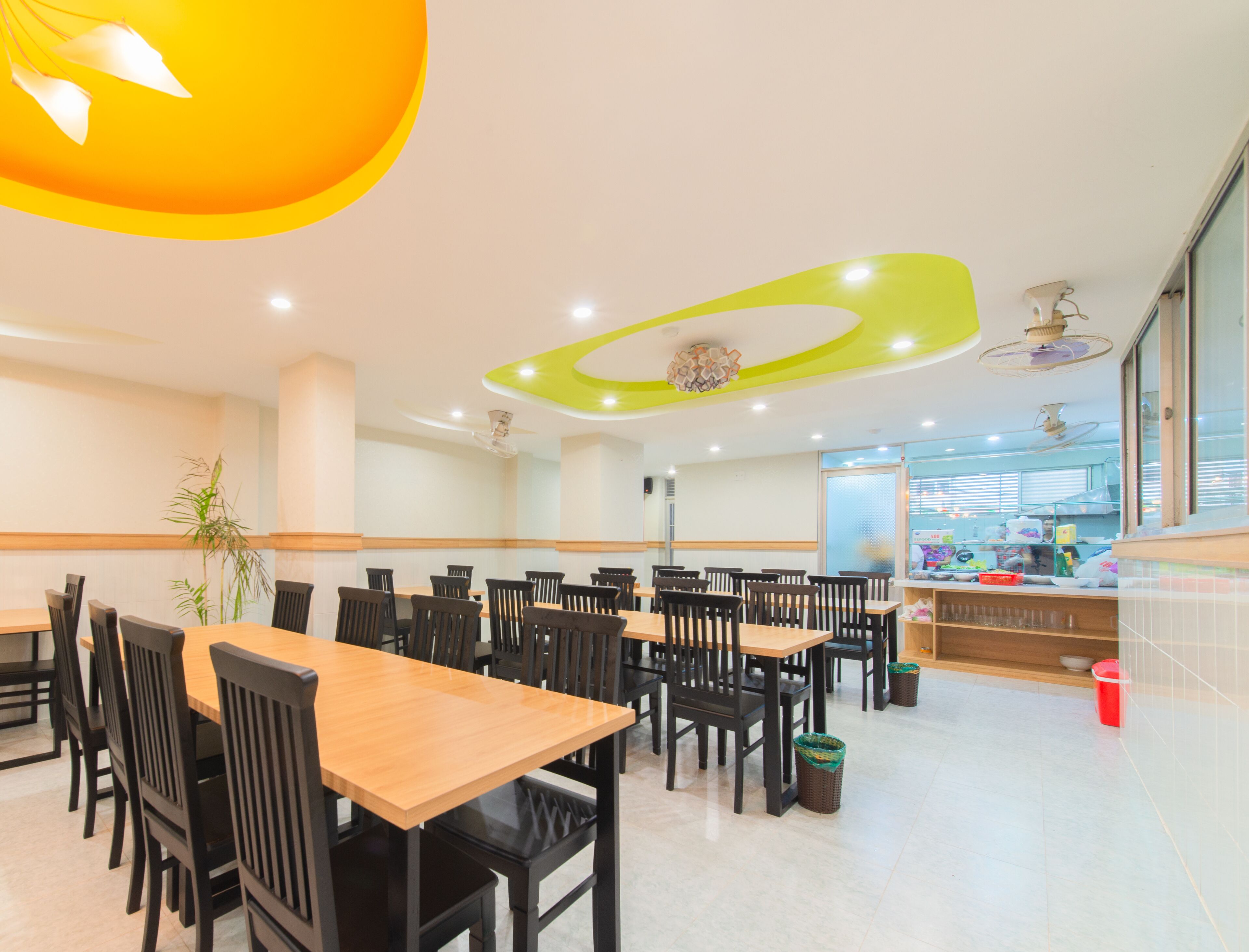 A modern dining area with wooden tables and black chairs, a central light fixture, and a view of the kitchen area in the background.